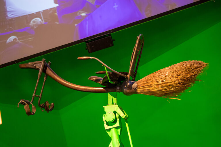 A long wooden broomstick with a bicycle-style seat, a backrest and foot stirrups, on a green stand in front of a green screen