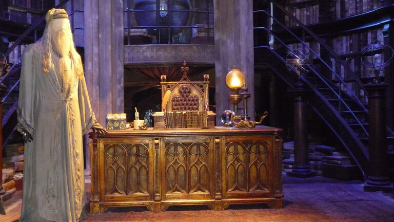 Dumbledore's long pale robes on a stand beside his carved wooden desk, with a lamp, a tall chair and spiral staircases up to shelves of books behind