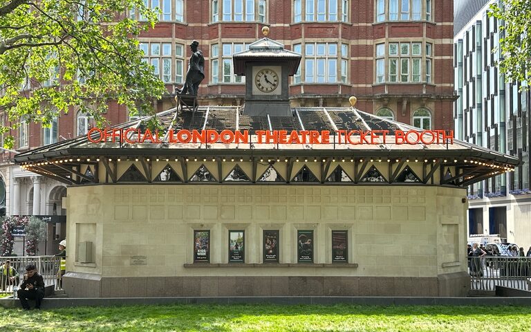 The Official London Theatre Ticket Booth in Leicester Square: a low stone kiosk with a clock tower and bright orange lettering along its roof, seen across the lawn