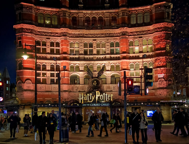 The red-brick Palace Theatre lit up at night, with a Harry Potter and the Cursed Child sign and gold wings over the entrance and crowds on the pavement