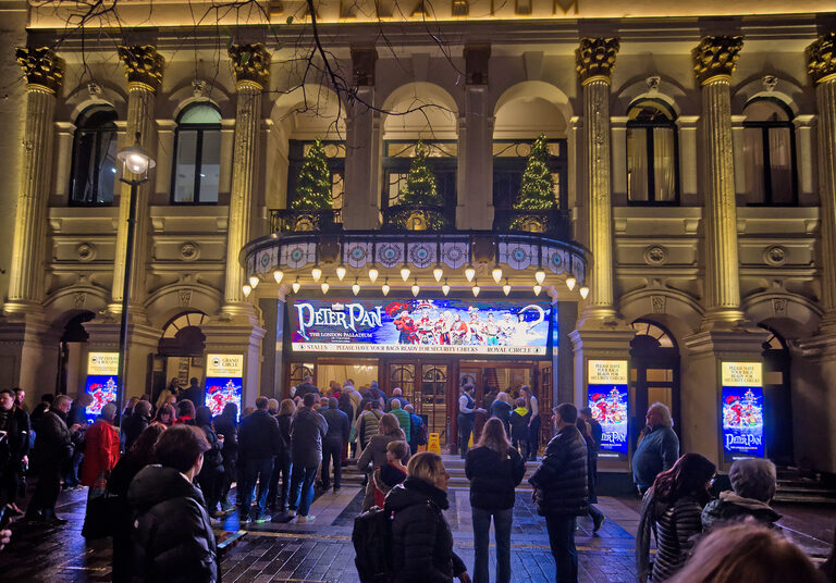 Crowds outside the London Palladium at night under a Peter Pan banner, with Christmas trees on the balcony above
