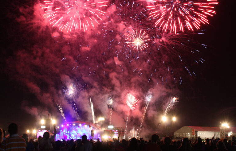 Fireworks bursting in pink and white over a lit-up stage, with a crowd in the foreground
