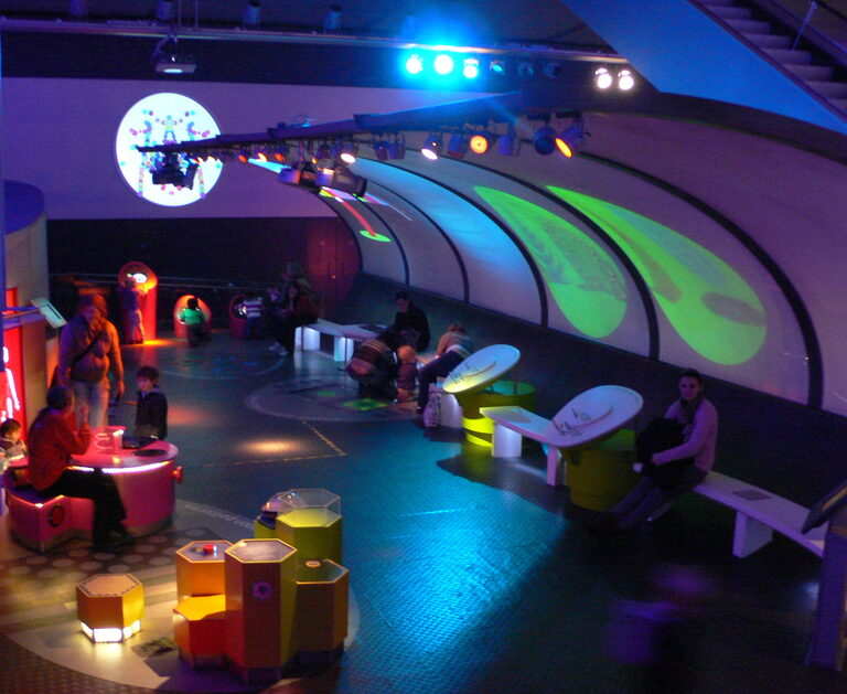 A dark room lit blue and green, with glowing seats and screens along a curved wall and children playing