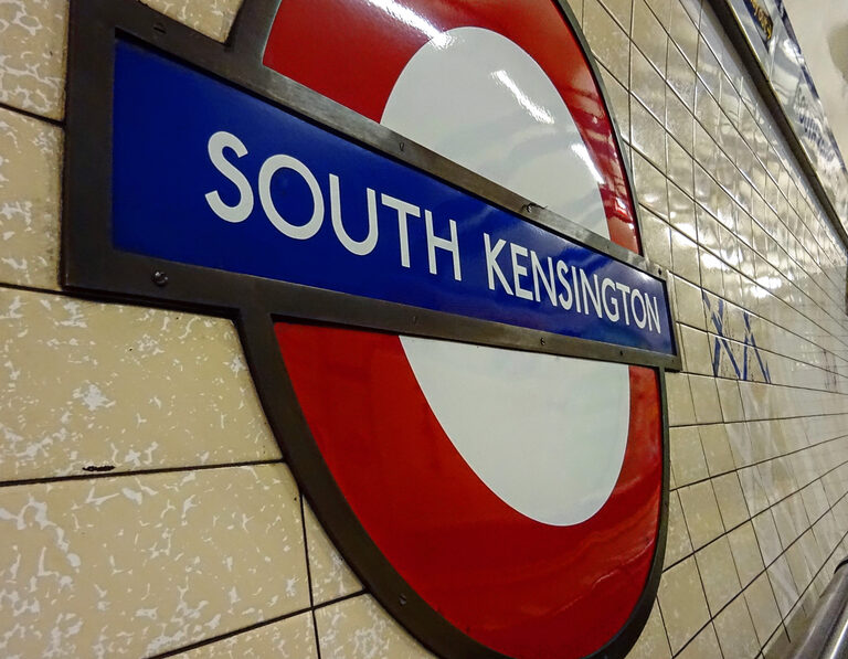 A red, white and blue Underground roundel reading South Kensington on a tiled platform wall