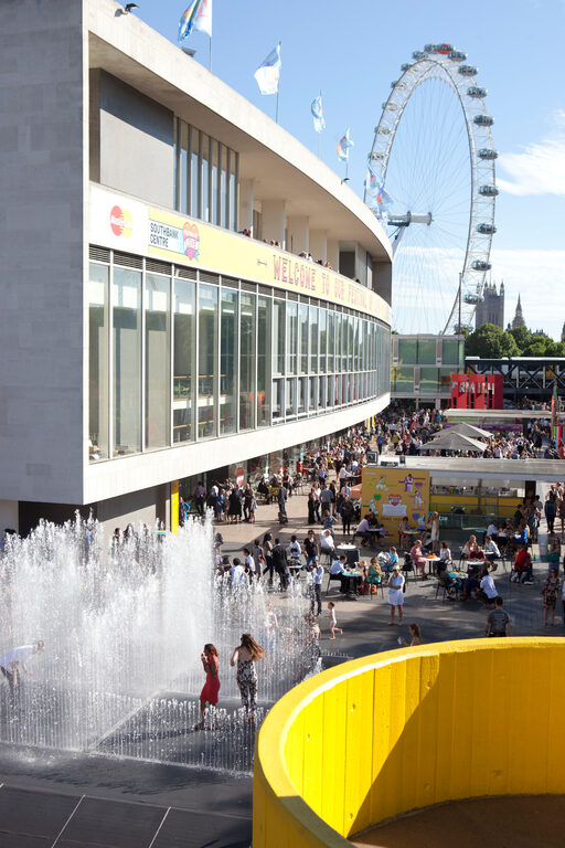 Children playing among jets of water in a fountain outside the Royal Festival Hall, with the London Eye behind