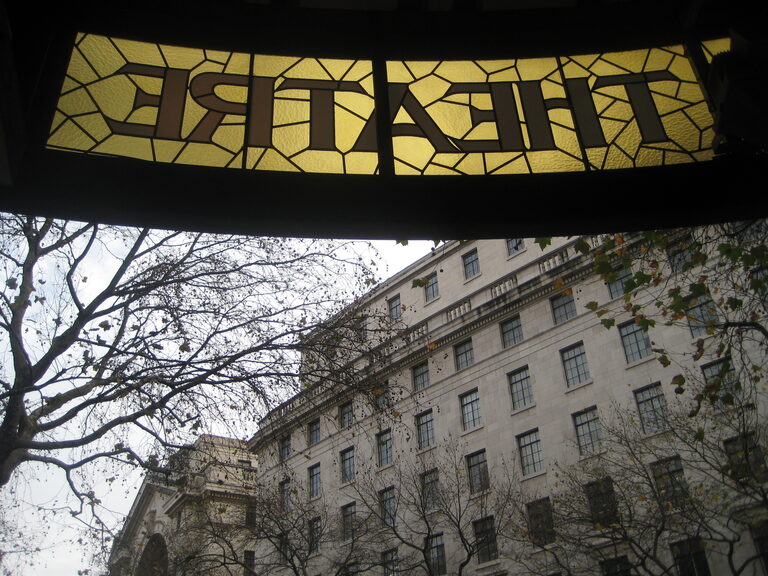 Looking up at a stained-glass THEATRE sign from underneath, with a white stone building and bare trees beyond